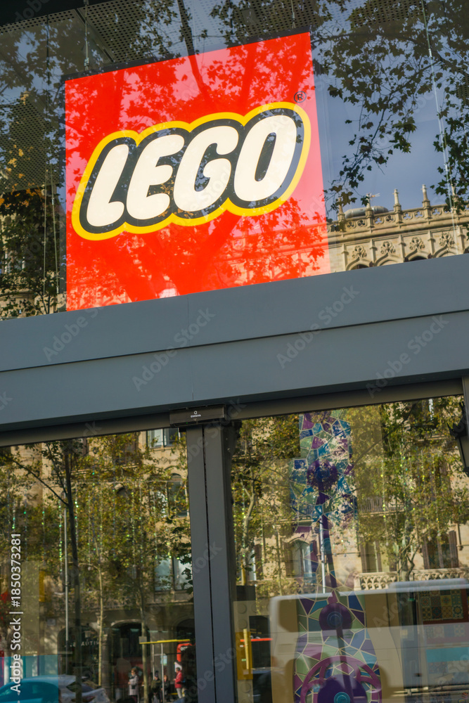 Fototapeta premium Barcelona, Spain, Europe - Nov 23, 2025: lego logo red yellow glass window storefront reflecting trees and historic architecture in a city street under blue sky with copy space for banner design
