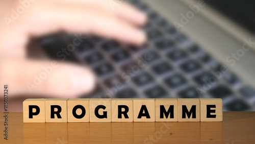 Handwritten text spelling out the word PROGRAMME on wooden blocks placed on a table, with a blurred finger poised over a keyboard in the background, reflecting a digital context.