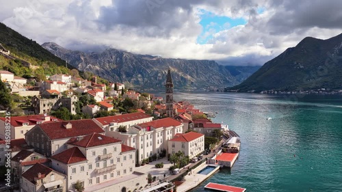 Aerial drone view of the coastal town of Perast, featuring stone buildings with red rooftops, a tall church bell tower, and the calm bay framed by rugged mountains under cloudy skies. Bay of