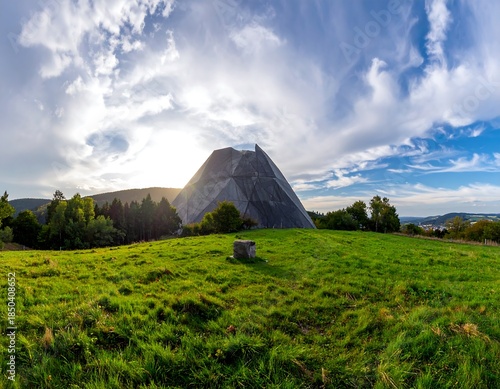 Pyramid-like structure on grassy hill under a bright sky with clouds and trees