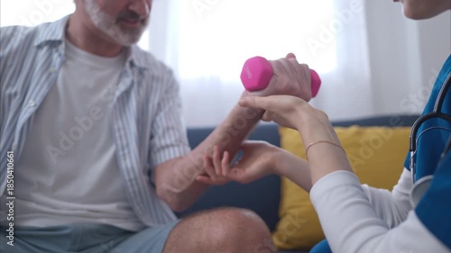 Female physiotherapist assisting an elderly man with arm strength training using a small dumbbell during home rehabilitation session