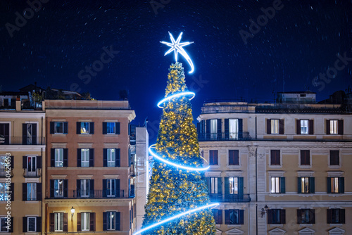 The Christmas tree decorated with yellow dot lights and decorated with white LED strips and a shooting star at the top. Christmas-themed nighttime cityscape.