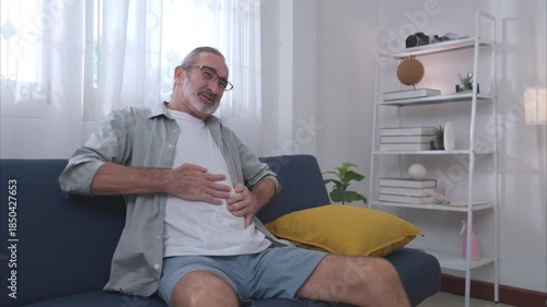 An elderly man sitting on a sofa holding his stomach in pain, illustrating symptoms of stomach pain from gastritis or a stomach inflame