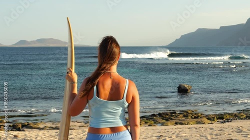 SLOW MOTION, CLOSE UP: Surfer with wooden surfboard watches waves breaking in offshore wind. Young woman is checking out empty surf spot on Lanzarote island before heading for a morning surf session.