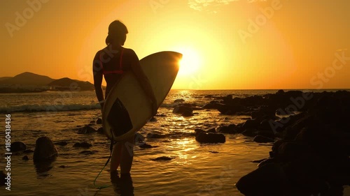 SILHOUETTE, LENS FLARE: Female surfer stands in shallow water, shielding her eyes as she looks out to sea at golden sunset. She is checking out the surf spot before heading for an evening session.