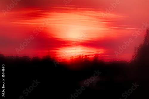 Fiery red sunset glowing above a dark treeline, with layered clouds creating a dramatic and atmospheric evening landscape.