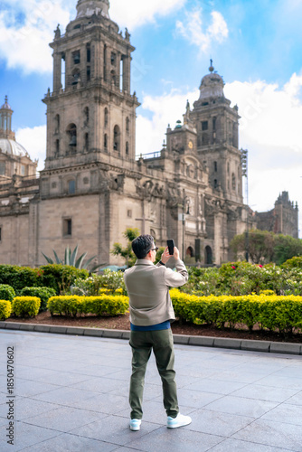 Unrecognized Latin man taking a photo of the Metropolitan Cathedral in Mexico City’s Zocalo, bright sunny afternoon, dressed in casual clothes, near green gardens, blue sky and historic architecture