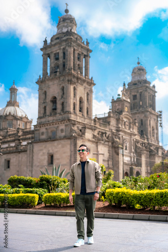 Young Latin man smiling and walking through the Zocalo of Mexico City during a sunny afternoon, dressed in casual modern clothes and sunglasses, with the grand Metropolitan Cathedral and open plaza