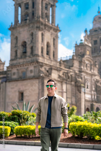 Young Latin man smiling and walking through the Zocalo of Mexico City during a sunny afternoon, dressed in casual modern clothes and sunglasses, with the grand Metropolitan Cathedral and open plaza