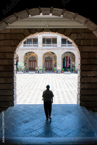 Unrecognized Latin woman walking the archway corridor of Museo Vivo del Muralismo in Mexico City, toward the courtyard adorned with murals and Mexican flags, surrounded by colonial stone architecture