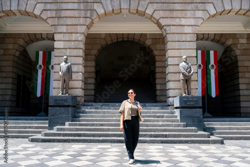 Smiling young Latin woman walking at courtyard of Museo Vivo del Muralismo in Mexico City, surrounded by statues, stone arches, and Mexican flags, wearing casual clothes and sunglasses a relaxed mood.