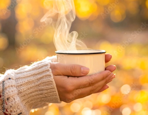 Close-up of hands holding a warm, white mug filled with steaming liquid. A blurred background of yellow and golden light