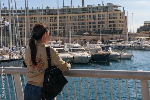 Turista mujer con mochila mirando hacia un puerto deportivo moderno lleno de yates y barcos de lujo frente a edificios residenciales en un día soleado de verano en el Mediterráneo.