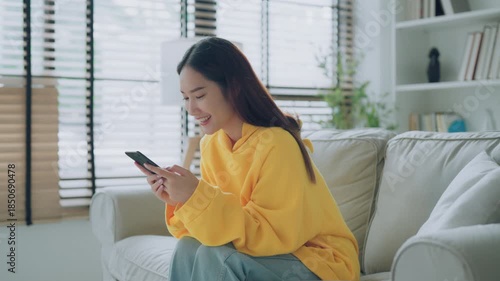 Happy young Asian woman relax sitting on couch using cell phone, smiling lady laughing holding smartphone, looking at cellphone enjoying doing online ecommerce shopping in mobile apps