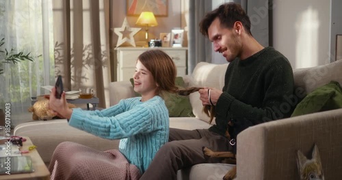 Father braids his daughter’s hair while the mother works remotely on a phone call. The girl takes a selfie, the mother joins them mid call at cozy couch. Happy modern family and work from home balance