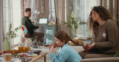 Father takes a break from working at home on laptop and approaches the mother braiding their daughter’s hair, kissing them in the living room. Family connection, comfort, and work from home lifestyle