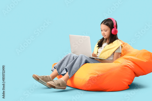 Young girl in headphones using laptop while sitting on bean bag chair on blue...