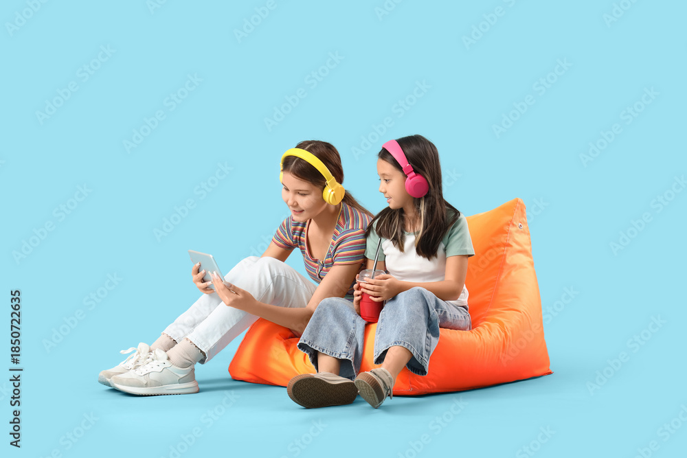 Obraz premium Cute Asian sisters in headphones with tablet computer watching cartoons on blue background