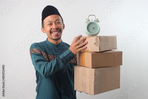Happy Asian Muslim man wearing a green traditional outfit and black songkok, smiling at the camera while holding a stack of cardboard boxes with Alarm Clock on Top. Isolated on a white background
