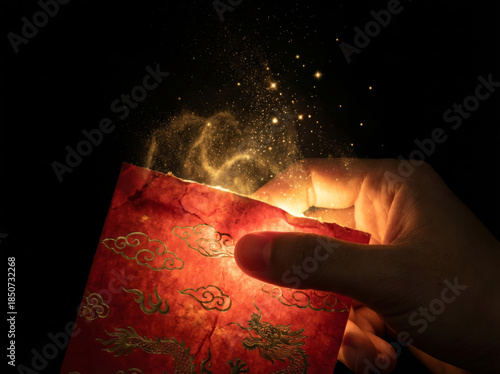 Macro shot of hand holding magical Chinese red envelope with rising golden dust and glowing sparkles against black background.