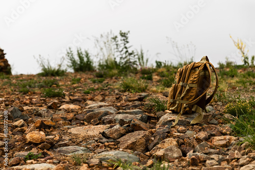 A green military backpack stands on a rocky mountain road in the grass, against a background of fog. The concept of military training, training camps, tactical exercises, and conscription.