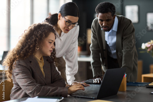 Young adult Caucasian woman sitting at desk using laptop while young Caucasian woman and Black man standing beside her, observing screen and discussing project