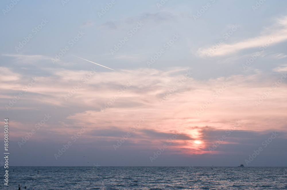 Fototapeta premium Pink, Cloudy sky at sunset; the trail of smoke from a jet aircraft is heading low to the right of the image.