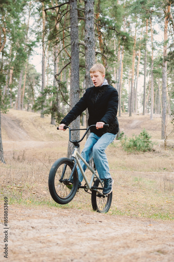 Fototapeta premium Young man performing a wheelie on a BMX bike in a pine forest, showcasing active lifestyle, youth culture, and extreme sports in nature. Dynamic outdoor activity.