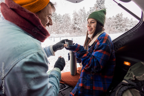 Couple taking a break while going for weekend vacation road trip, drinking hot tea in a car trunk