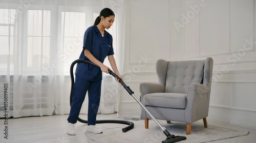Wallpaper Mural A person wearing a blue uniform is vacuum cleaning a room. They are standing behind a light grey armchair and holding the vacuum cleaner with both hands Torontodigital.ca