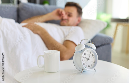 Photography Man lying on sofa under white blanket near alarm clock in the morning at home waking up