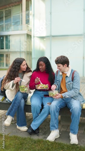 Group of multiethnic university students eating a healthy lunch together sitting on a campus bench
