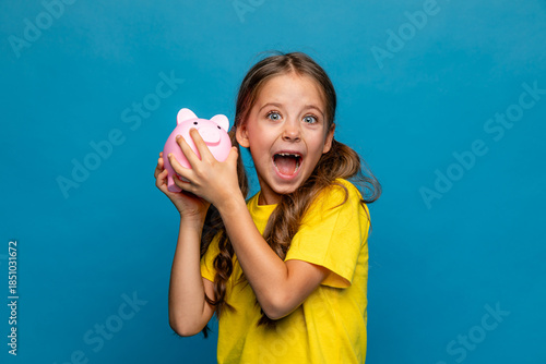 Funny little girl in yellow t-shirt holding pink piggy bank money box and looking at camera on blue background. Financial concept of children pocket money. Save money for education and family expenses