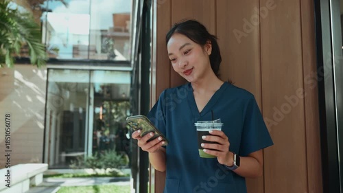 Smiling young asian female doctor in scrubs taking a break outdoors. She is happily using her smartphone while holding a cup of coffee