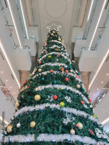 Low angle view of a giant decorated Christmas tree in a modern shopping mall atrium. Festive holiday decorations with baubles, tinsel, and a star topper under bright interior architectural lighting