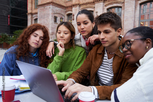 Multiracial group of students gathering to study at university campus, concentrated looking at a laptop together, working on a school project as a teamwork. Copy space.