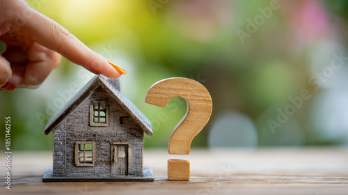 Hand touching a miniature model house placed beside a wooden question mark symbolizing uncertainty or questions about home ownership or real estate decisions on a bl