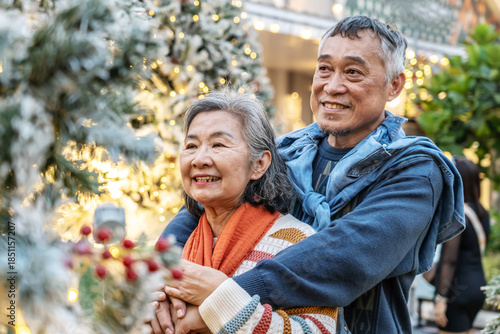 Elderly asian couple embraces near a beautifully decorated Christmas tree. The husband places a warm scarf around his wife, filling the air with love and festive cheer as they share joyful smiles.
