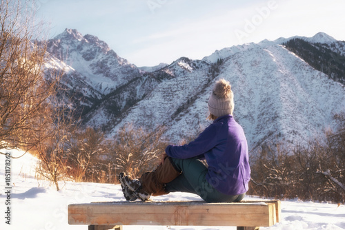 A woman sits on a wooden bench, enjoying a sunny winter day in the mountains. Rear view. Snowy landscape, bright sun, serenity, outdoor recreation, hiking, hiking, Alatau mountains