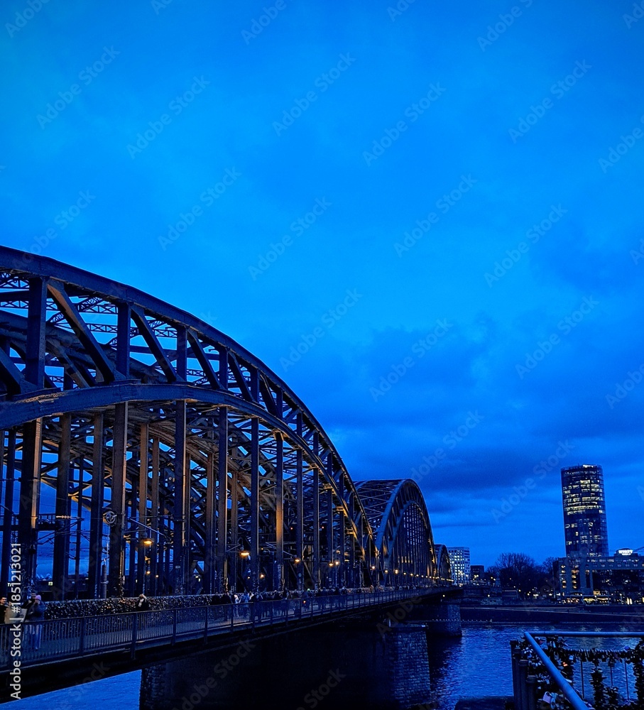 Fototapeta premium Evening landscape, city across the Rhine River in Cologne, Germany, December 19, 2025. Evening city on the riverbank. Reflection in the water. Night blue sky. 