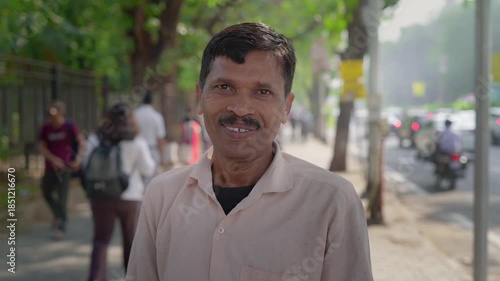 A middle-aged Indian Asian happy middle aged man or male standing outside in the daylight by a busy city road on a sidewalk, looking at the camera and smiling, with pedestrians walking in background