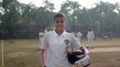 An Indian young, aspiring girl or female cricketer in a jersey and holding a helmet, stands outdoors on the cricket ground, smiles facing the camera, with teammates preparing in the background