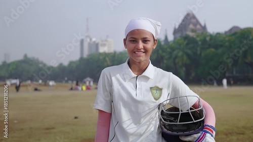 A happy Indian young, aspiring girl or female cricketer in a jersey and holding a helmet, prepared to go out to bat on the field, stands outdoors on the cricket ground, smiles, facing the camera, 