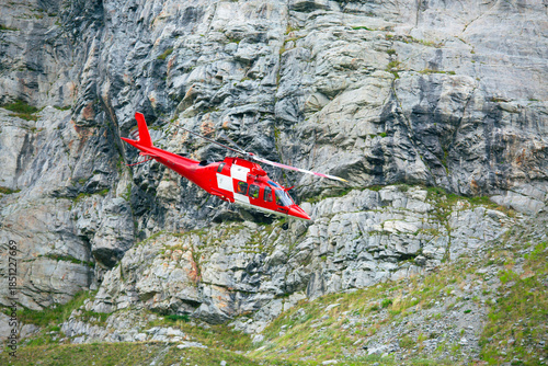 Red rescue helicopter flying along steep rocky mountain wall
