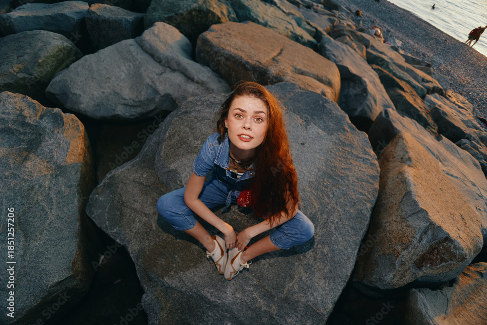 Fototapeta premium young woman sitting on rocks near water wearing denim overalls with long red hair and relaxed expression at sunset outdoors nature scene