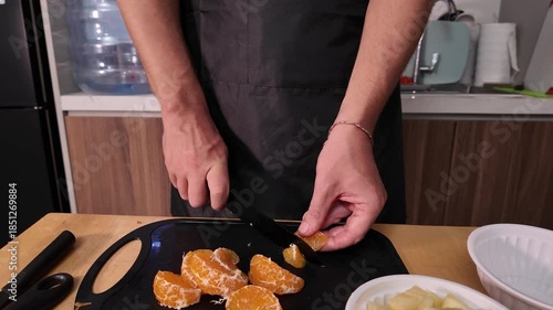 The hands of a man in a apron who is cutting a peeled mandarin into slices.