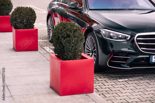 A black car is parked on a brick road near a sidewalk with red square planters containing round bushes. The scene is in a modern, urban setting.