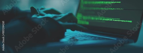 Close-up of a person's hands typing on a laptop keyboard with green coding on the screen in a dark room with blue lighting.