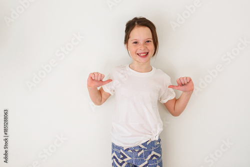 Portrait of a smiling girl standing in front of a white wall pointing her thumbs at herself