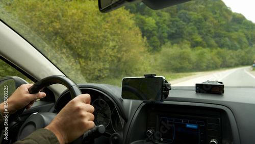 Autotourism. A car cabin with a navigation app on a smartphone. The driver's hands are on the steering wheel. A mountain road is visible outside the windshield.